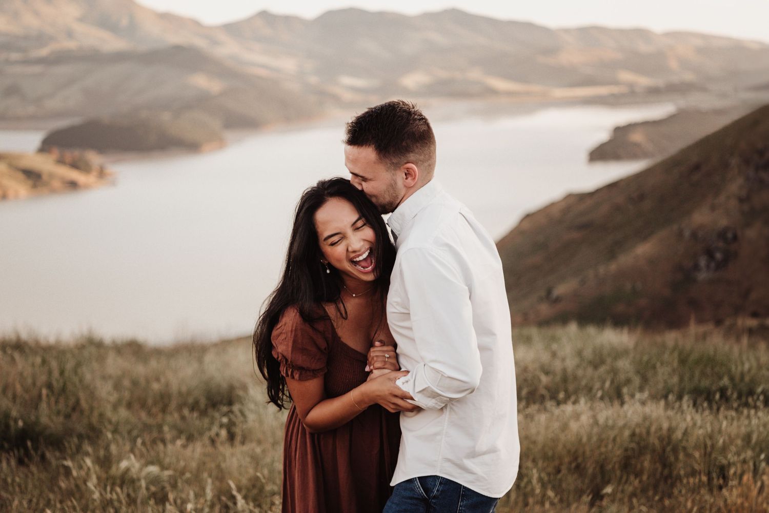 A couple shares a joyful moment together on a hilltop overlooking a serene mountain lake during golden hour.