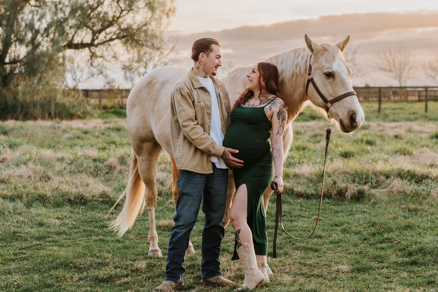 A couple stands in a golden sunset field with their white horse on a rural farm setting.