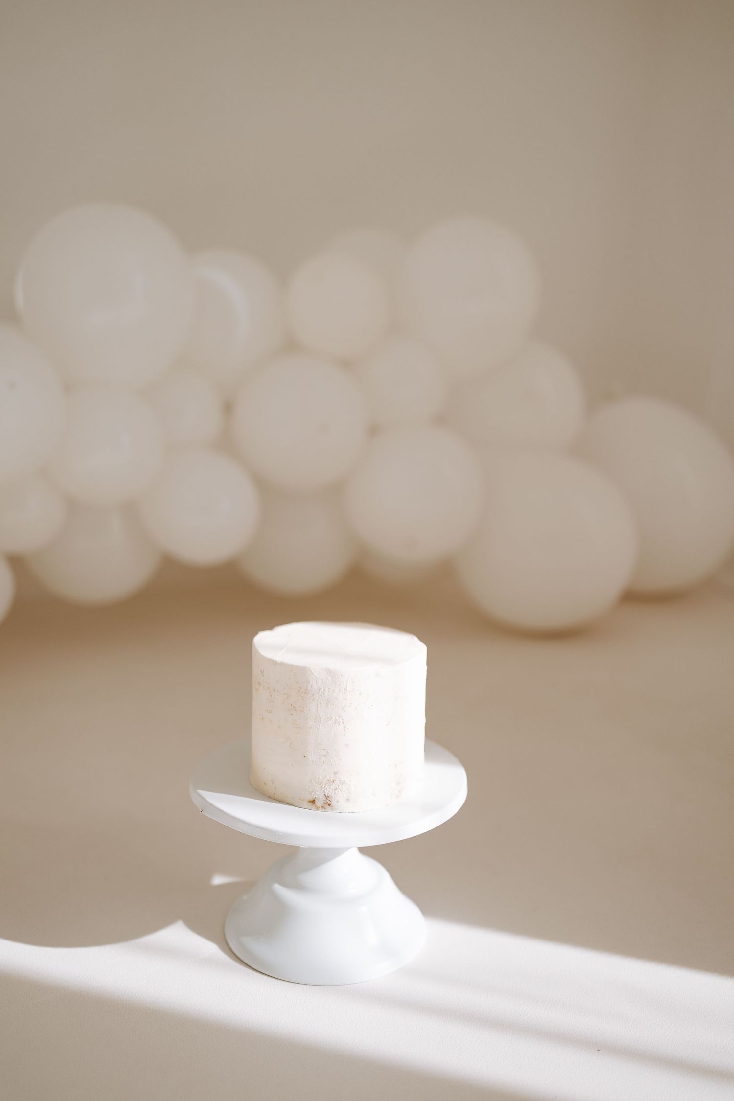 A minimalist birthday cake with a number one candle sits atop a white cake stand on a wooden surface.