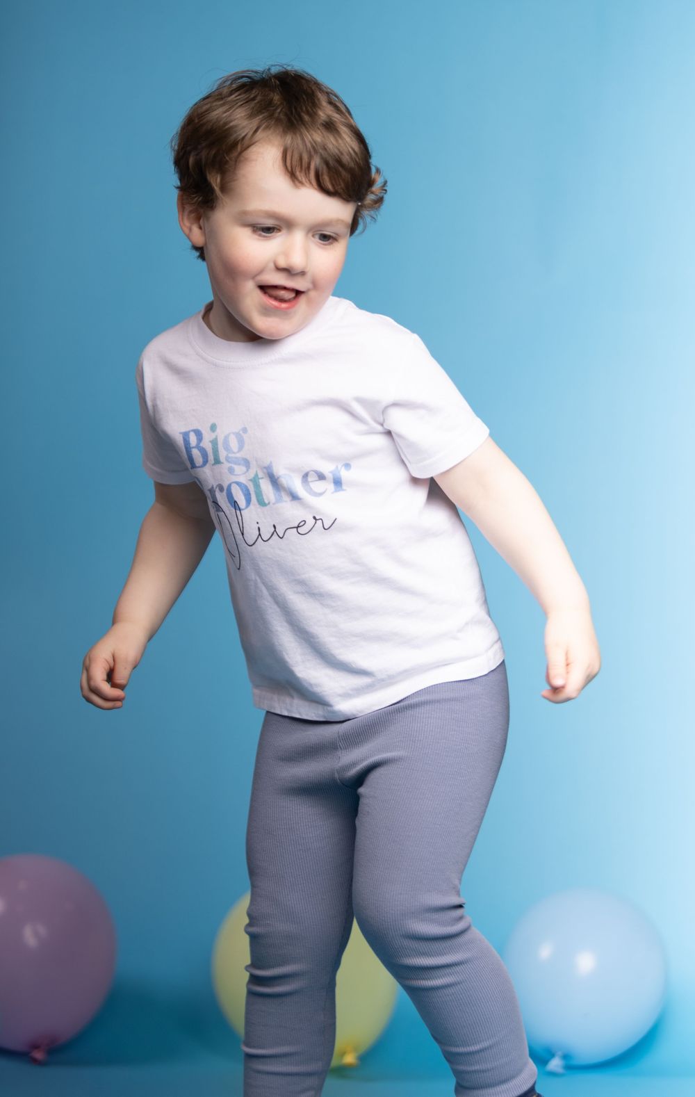 A child in a white t-shirt and gray leggings dances playfully against a blue background with colorful balloons.