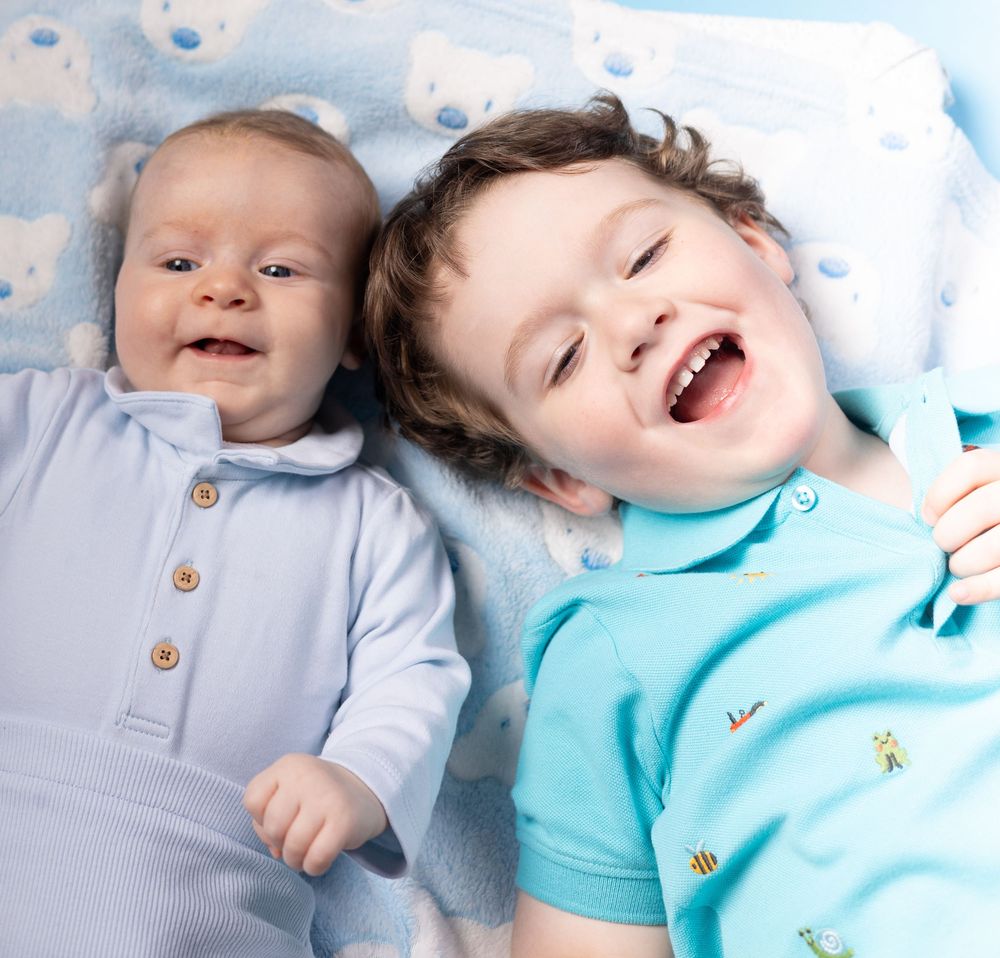 Baby in gray and turquoise outfits laying on white bedding while smiling and laughing.