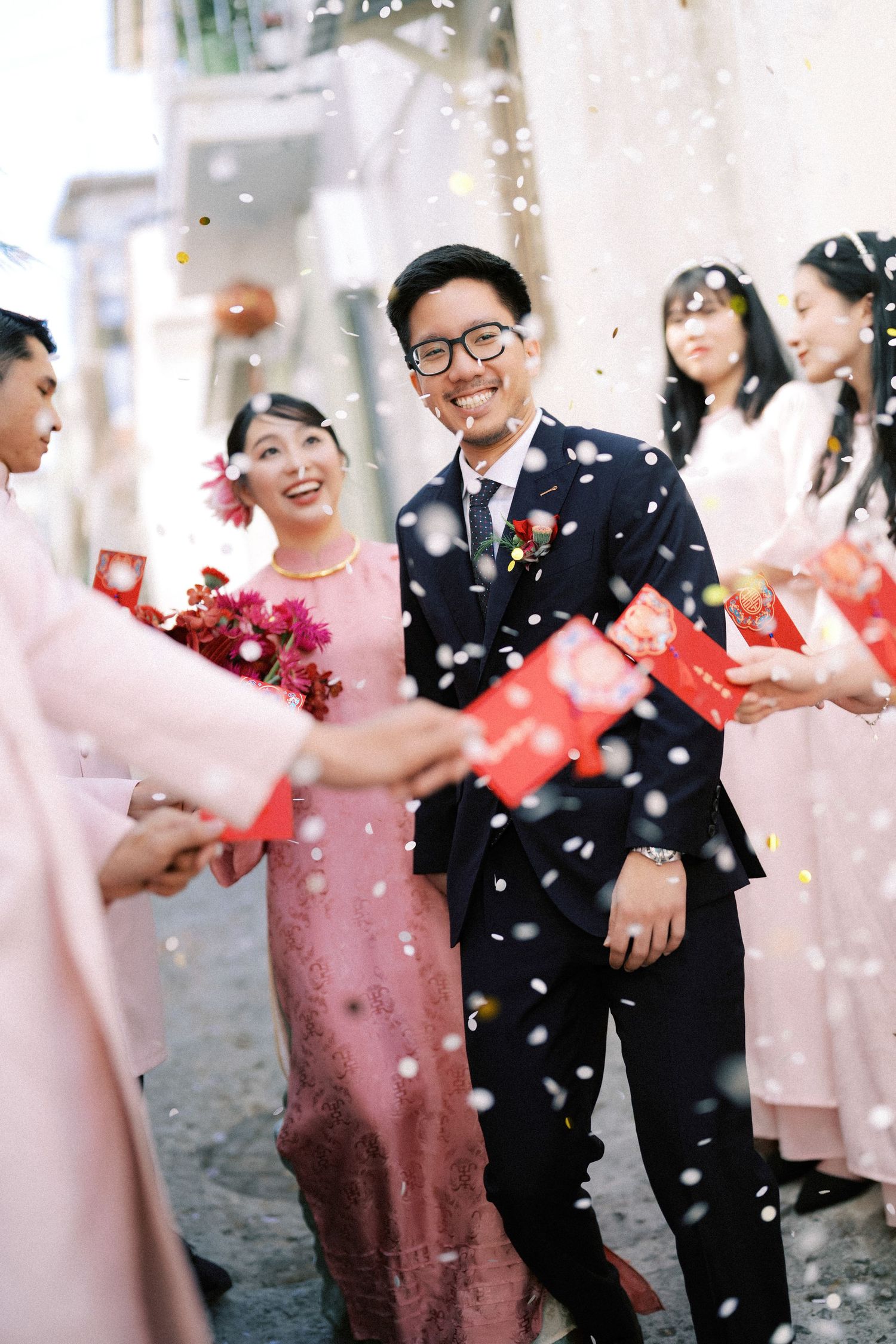 Wedding celebration in pink and black attire with guests exchanging red envelopes amid falling confetti.