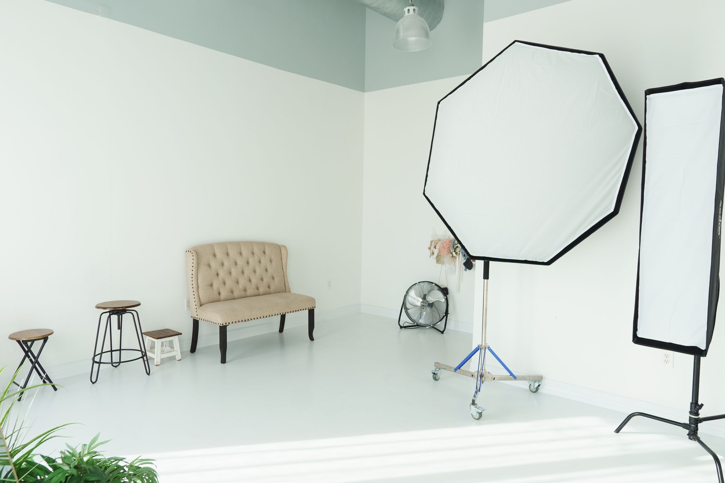 Professional photography studio setup with white walls, lighting equipment, and tufted seating furniture.