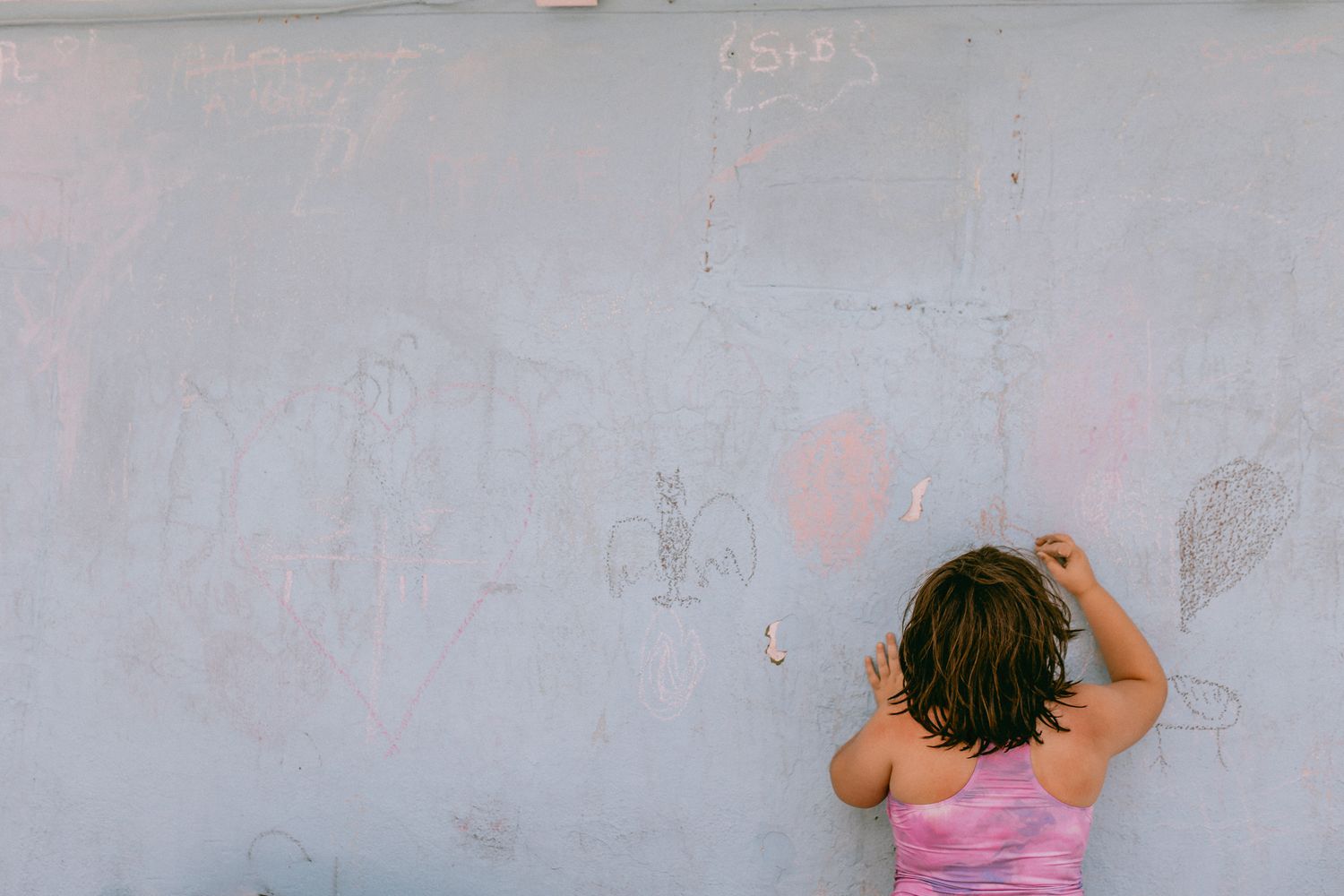 A child with dark hair wearing a pink top stands facing a white wall with faint markings drawing with chalk. Whitney Popp, Popp Photography