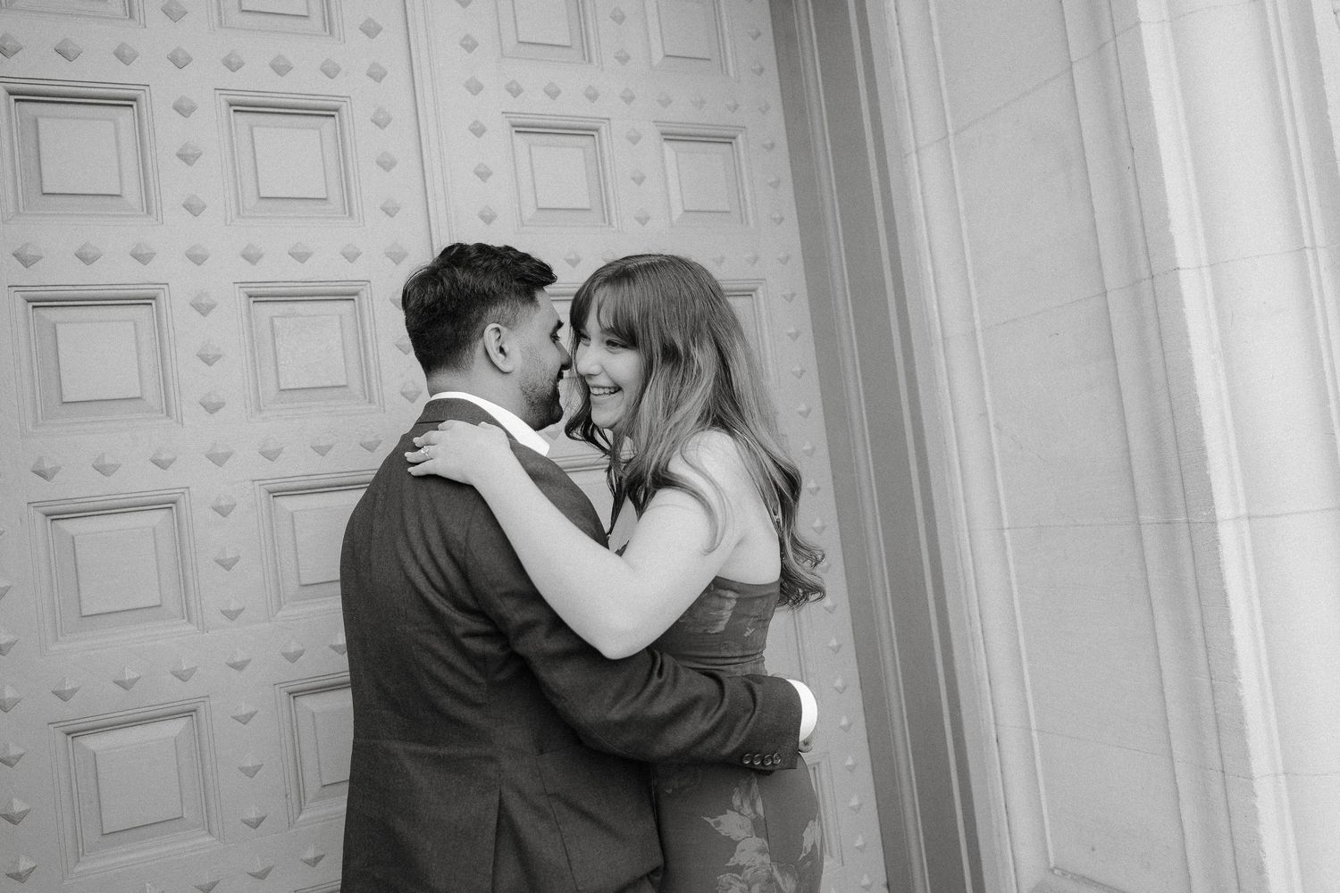 A romantic couple embraces in front of a white paneled wall in a black and white photograph.