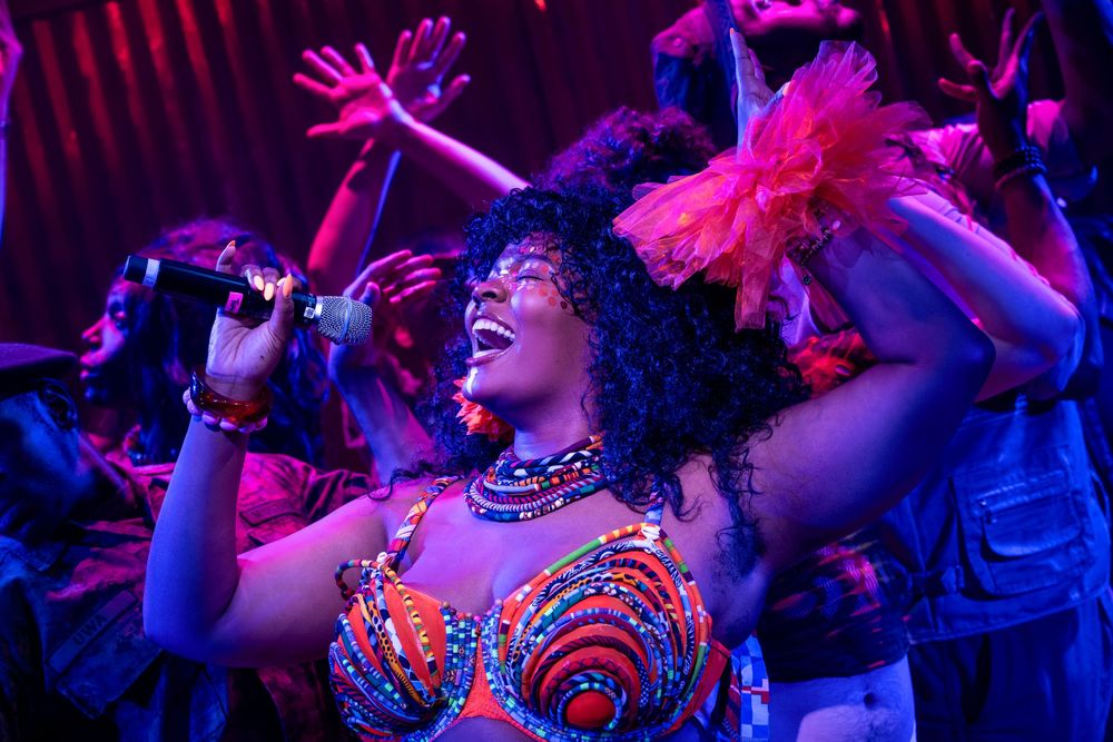 A woman in colorful African print performs on stage with a microphone under vibrant purple and red lights.