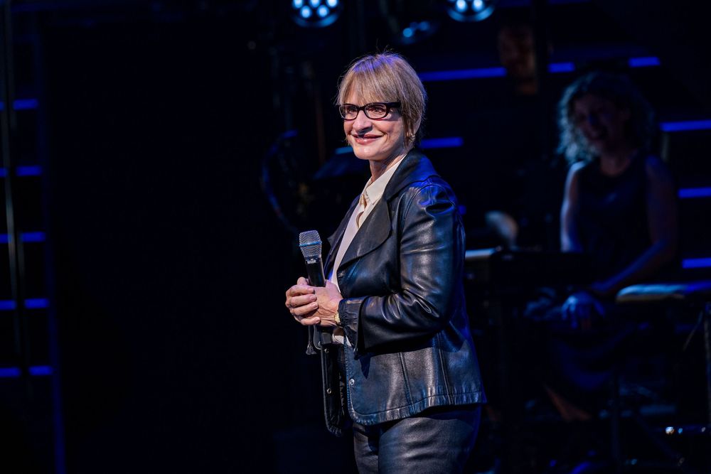 Woman in silver jacket holding microphone on stage performing under blue spotlight at live music event.
