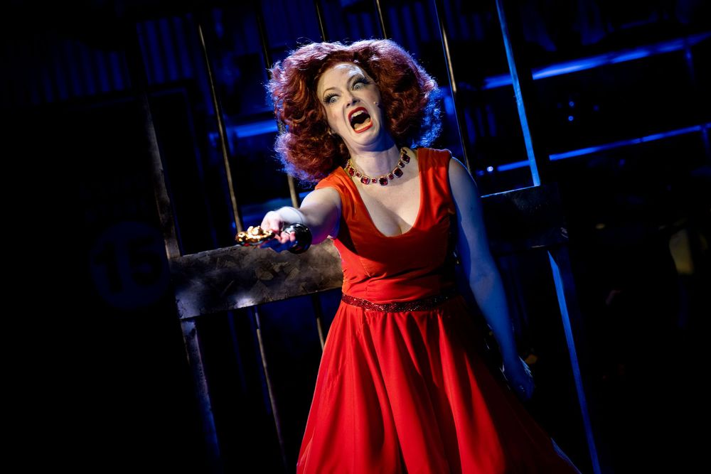 Actress in red dress performing on stage, singing dramatically under blue spotlight in theatrical production.