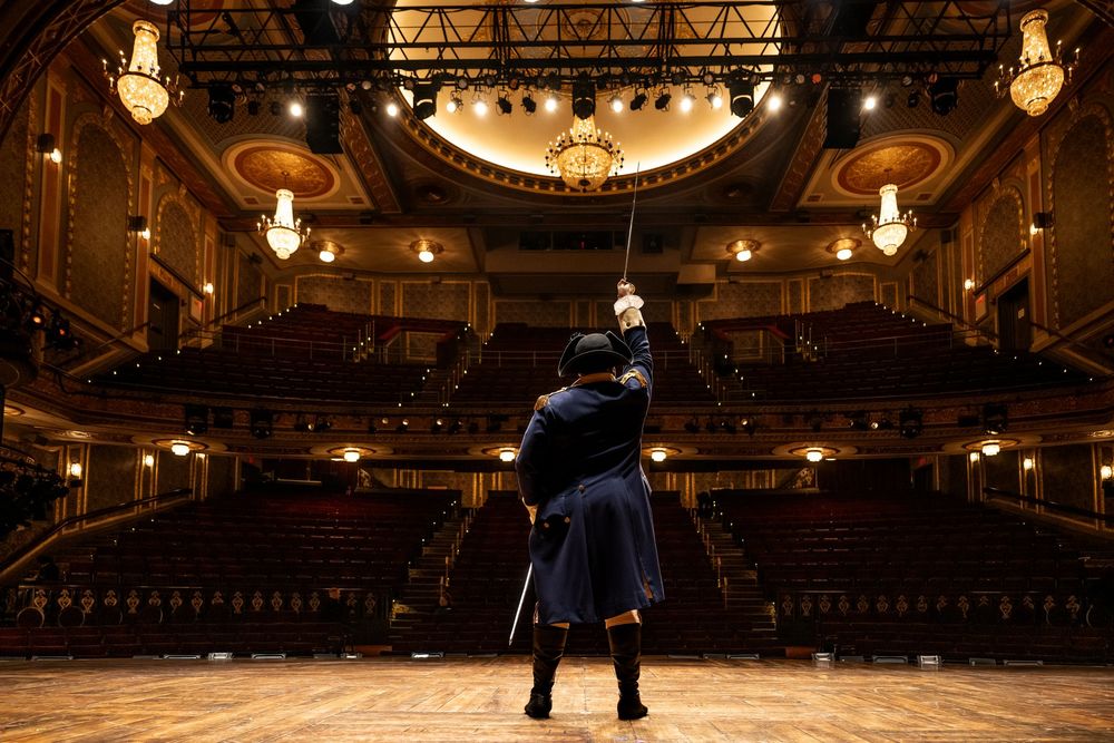 A couple dances gracefully in an elegant ballroom under warm lighting and ornate chandeliers.