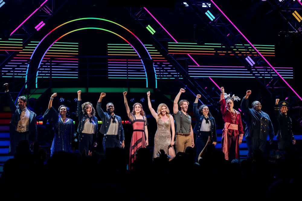 Broadway cast takes a bow on stage with colorful rainbow arch lighting during a musical theater curtain call.
