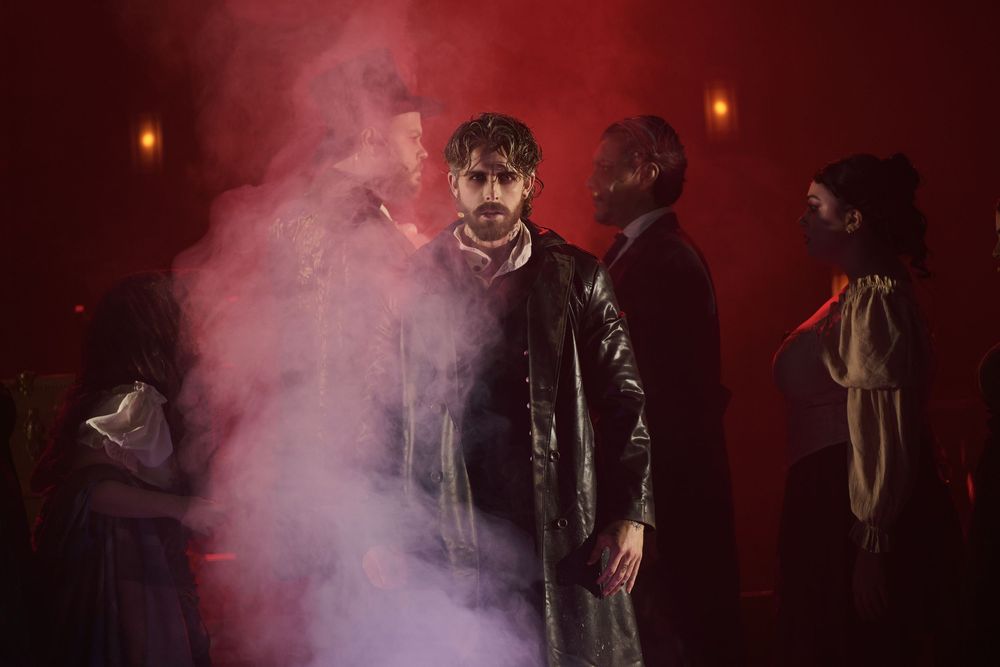 Dramatic theater scene with actors in dark costumes surrounded by red smoke and moody lighting on stage.