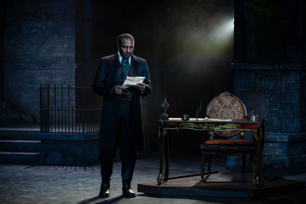 A man in a dark coat reads a letter under a spotlight on a dramatic stage set with a ornate desk.