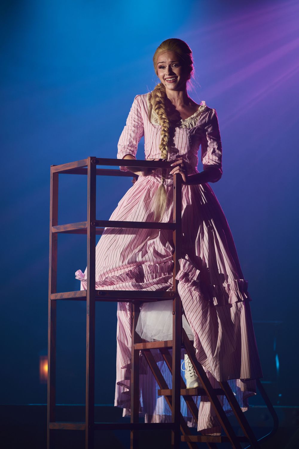 Woman in pink Victorian dress smiling on wooden platform under blue stage lights.