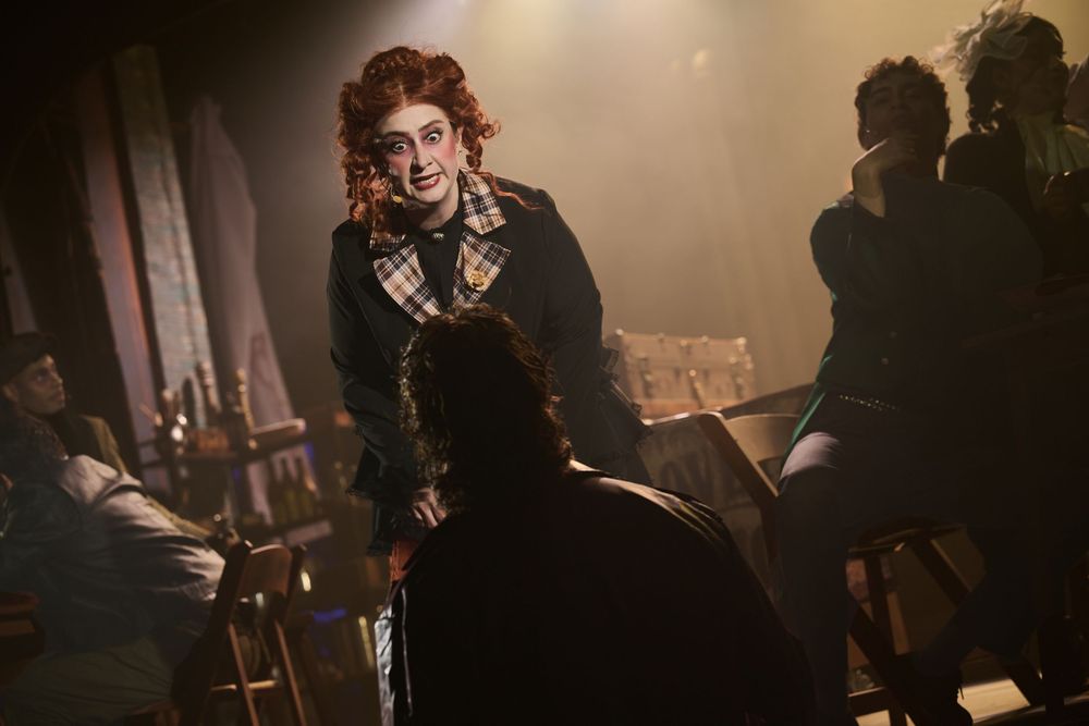 Actor in Victorian costume with curly red hair performs on a dimly lit theatrical stage.