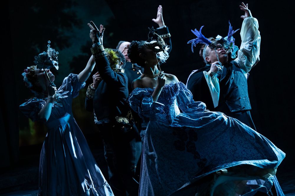 Performers in blue masquerade costumes dance dramatically on a dark stage with raised arms and flowing gowns.