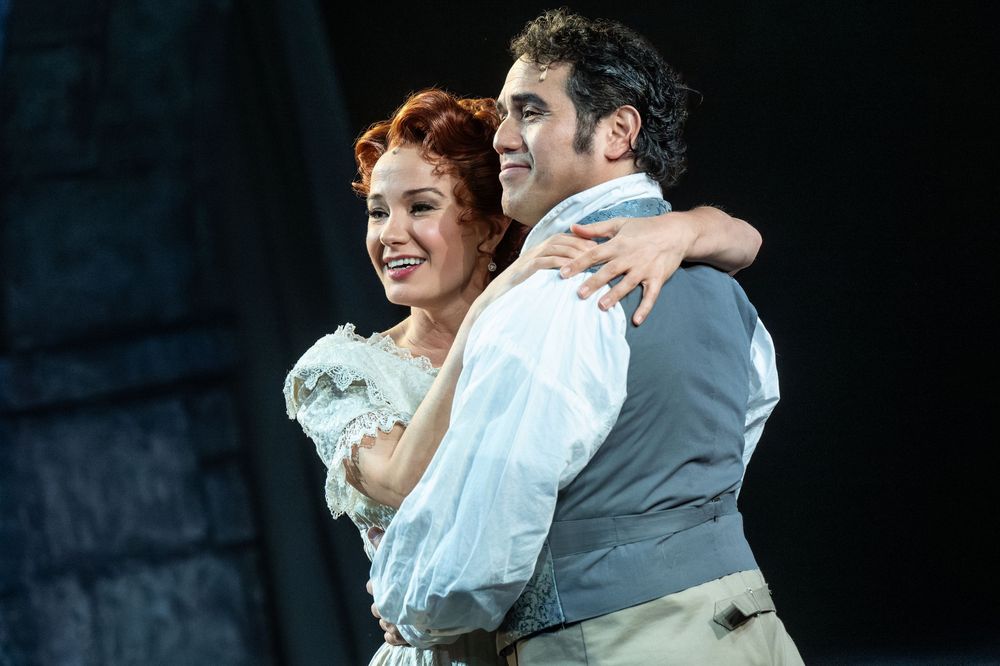 Two performers embrace on stage in period costumes during a musical theater production.