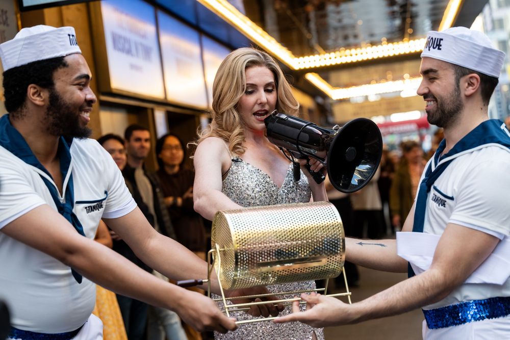 Woman in silver gown uses megaphone flanked by two men in sailor costumes holding a gold drum outdoors.