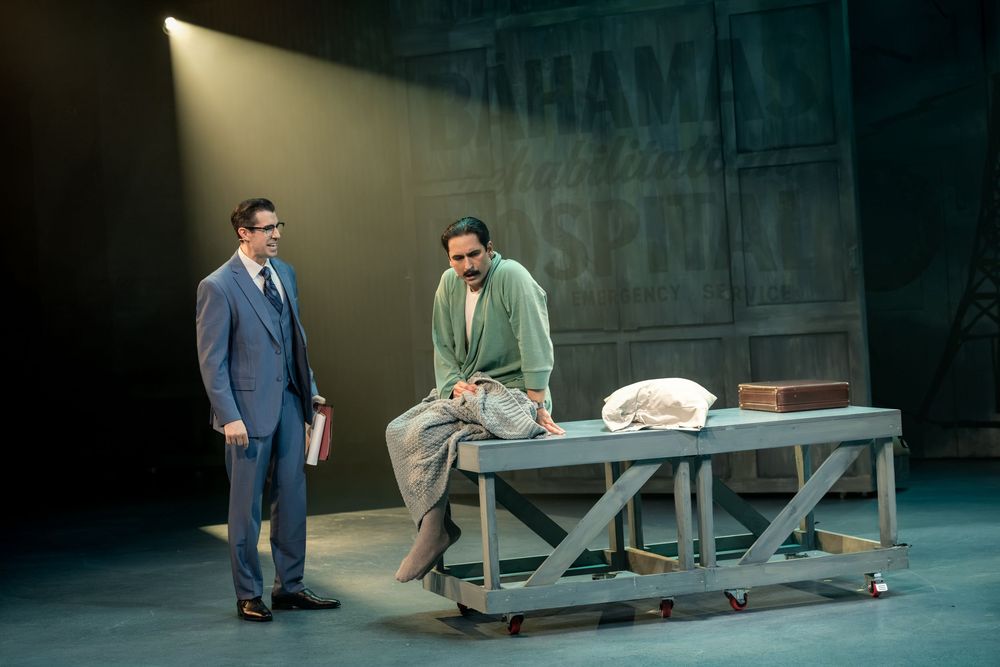 Two actors perform a dramatic theater scene, one in a suit and one in a robe, on a minimalist stage with moody lighting.