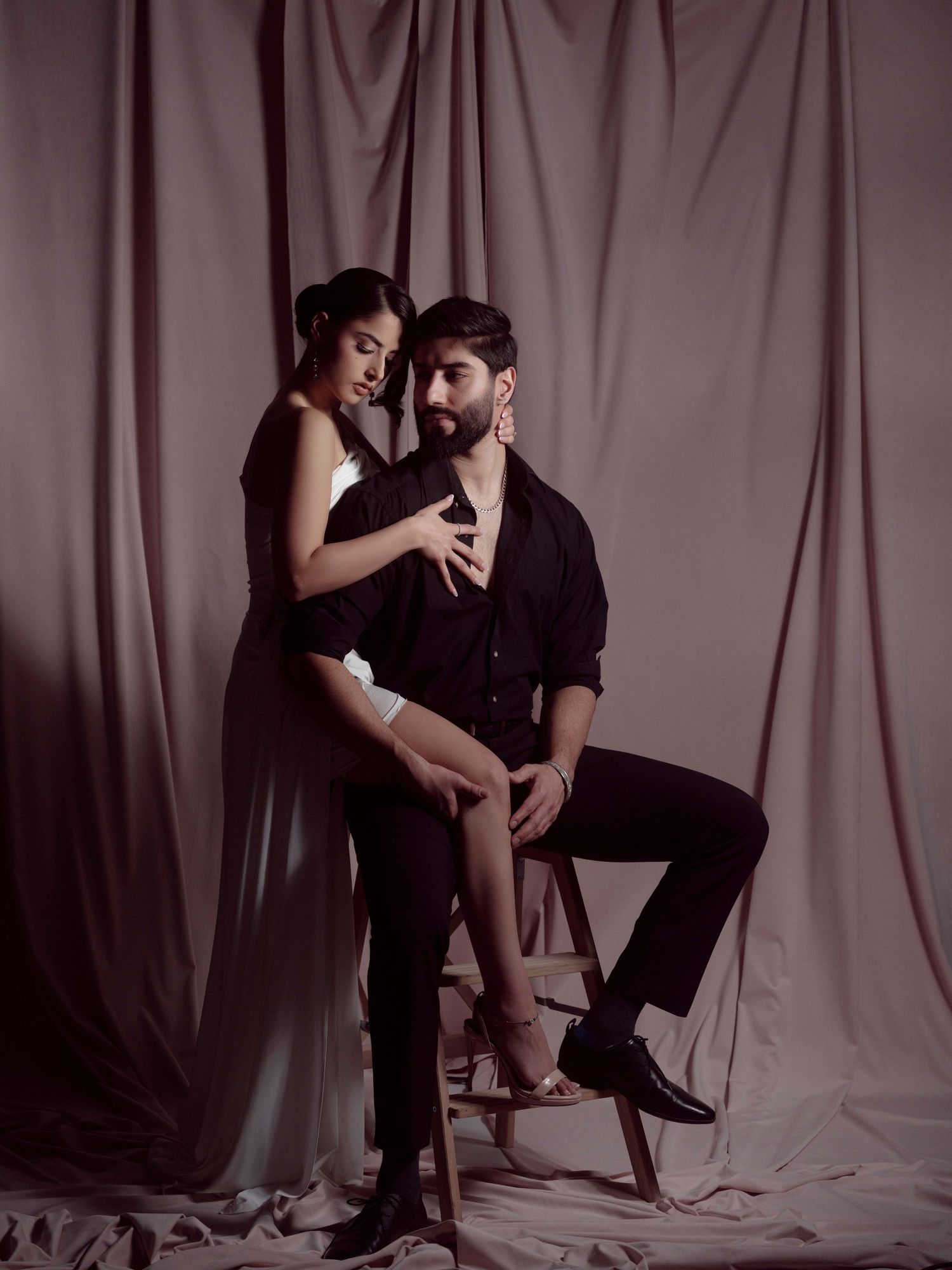 Dramatic fashion portrait of a couple in dark attire posing intimately against draped fabric backdrop in moody lighting.