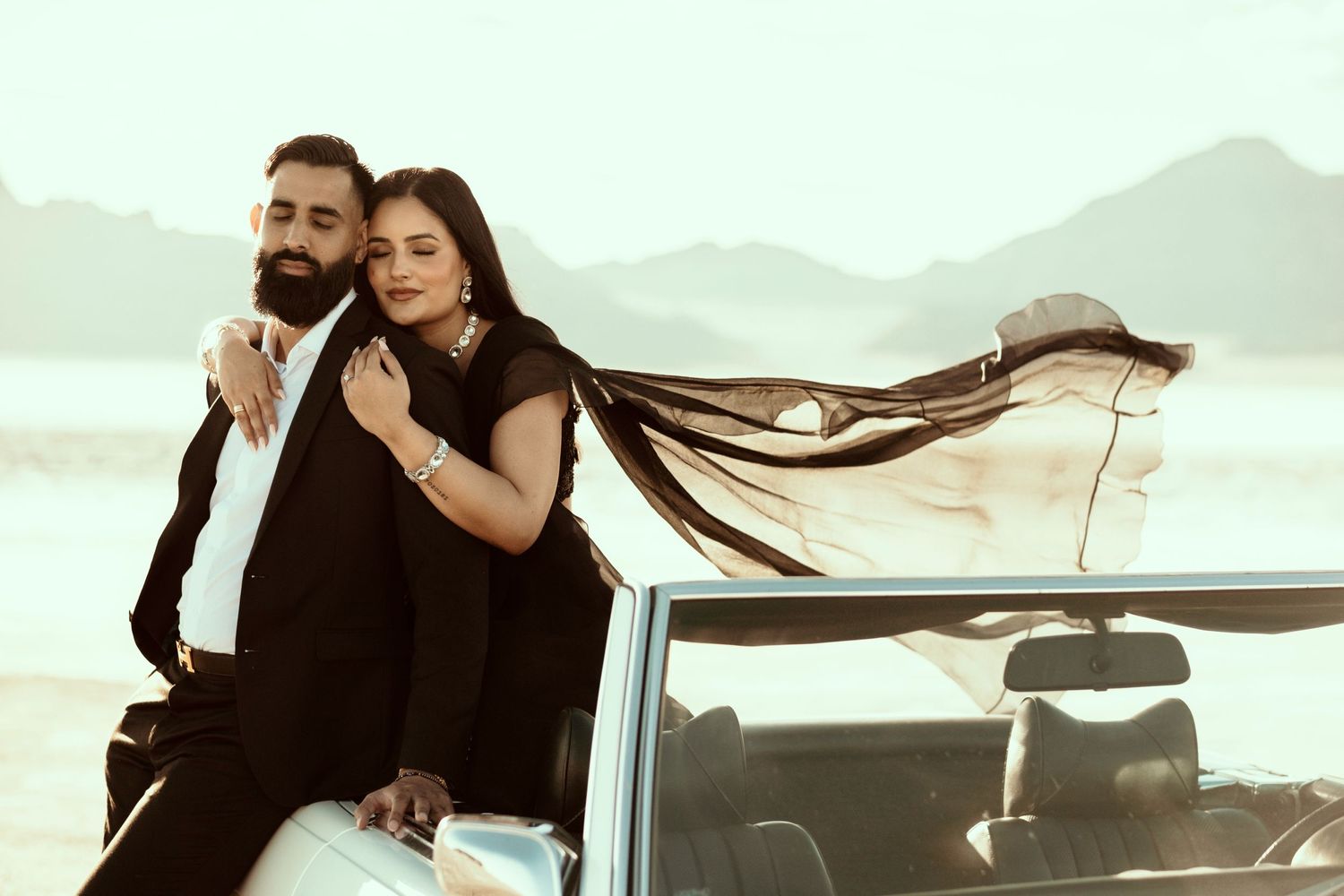 Couple in black outfits pose together against mountain backdrop as wind blows long hair by convertible car.