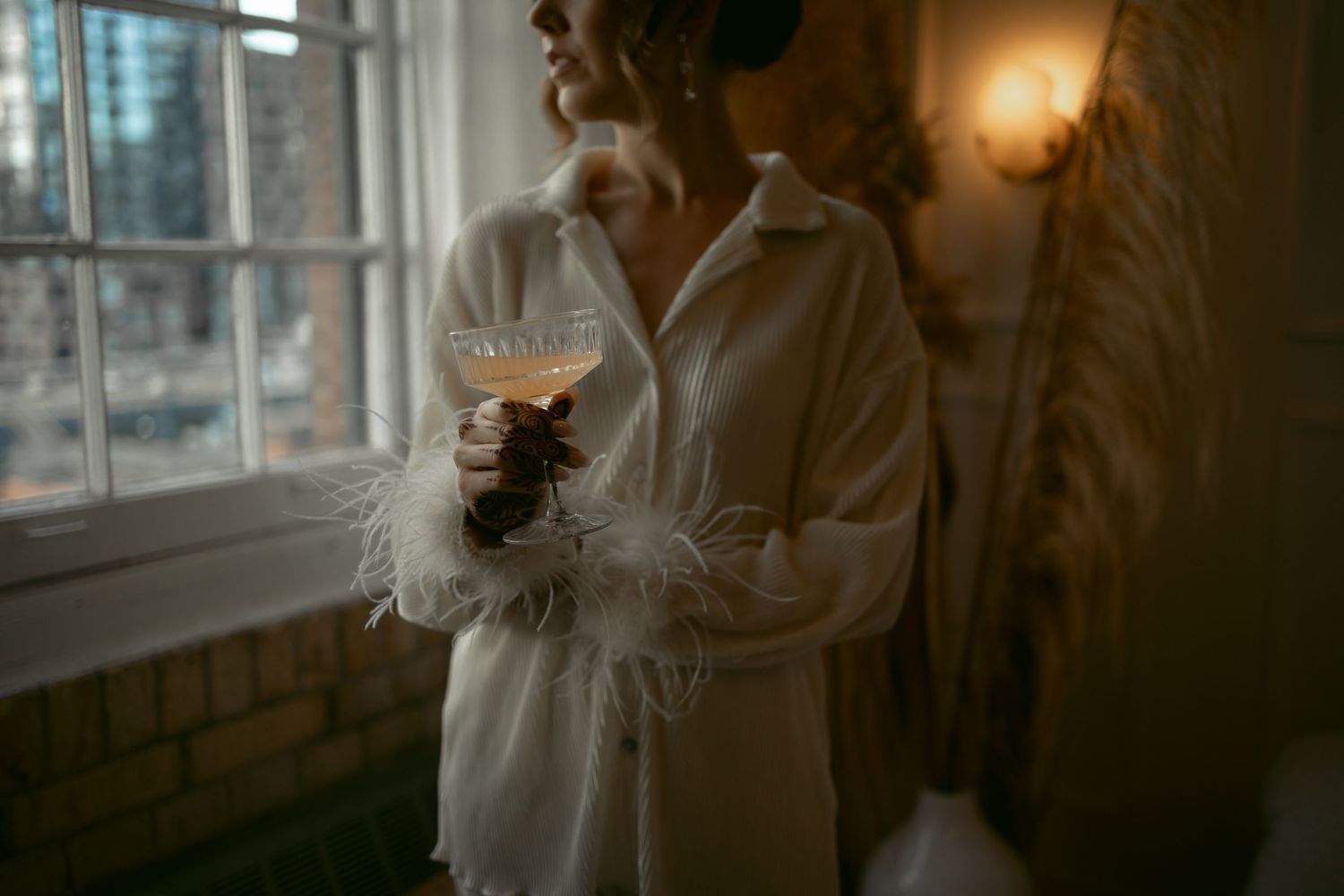 Someone in a white robe holding a wine glass stands by a window in a dimly lit room creating a cozy atmosphere.