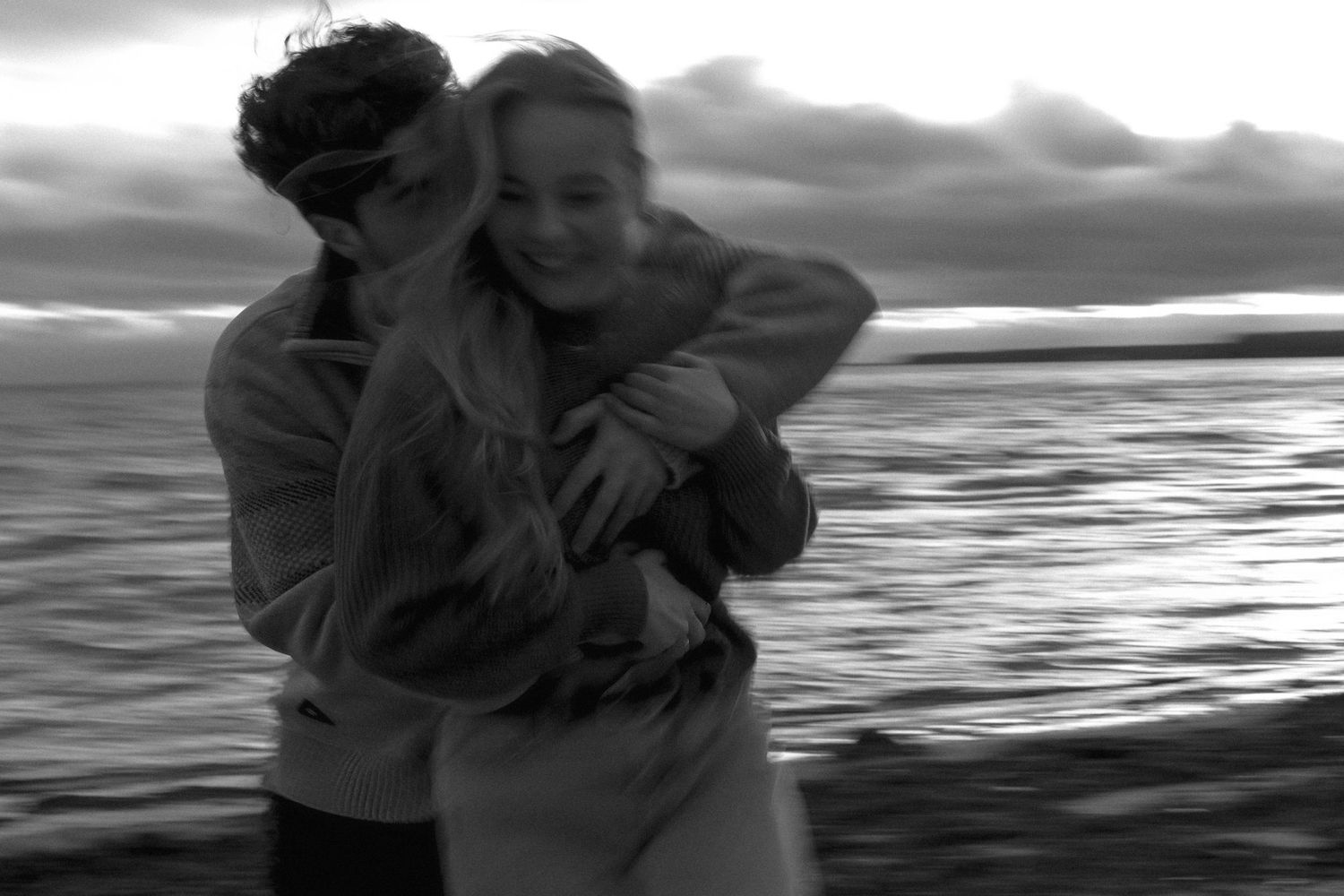 A black and white photo of two people embracing by a body of water at sunset with dramatic cloudy skies.
