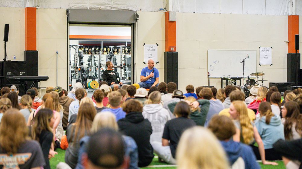 Large crowd gathered in an indoor venue with orange pillars and white walls for a presentation.