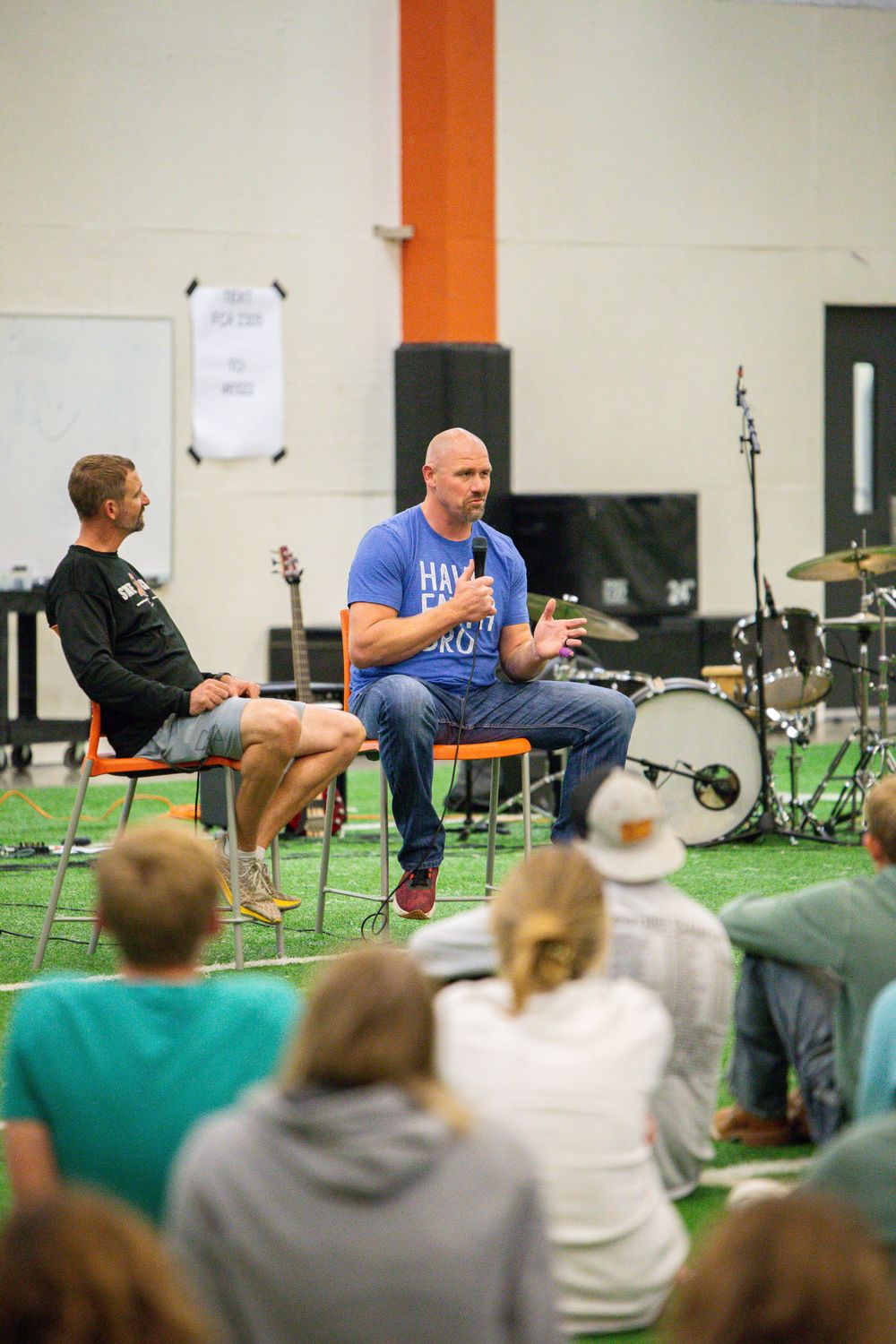 Audience members seated in chairs watch presenters on a stage with green turf flooring in an indoor venue.