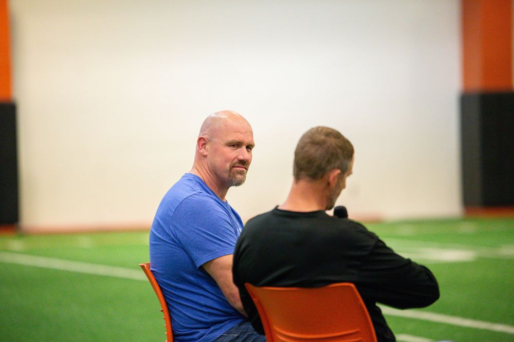 Presenters seated in orange chairs on stage during an indoor event with green turf flooring.