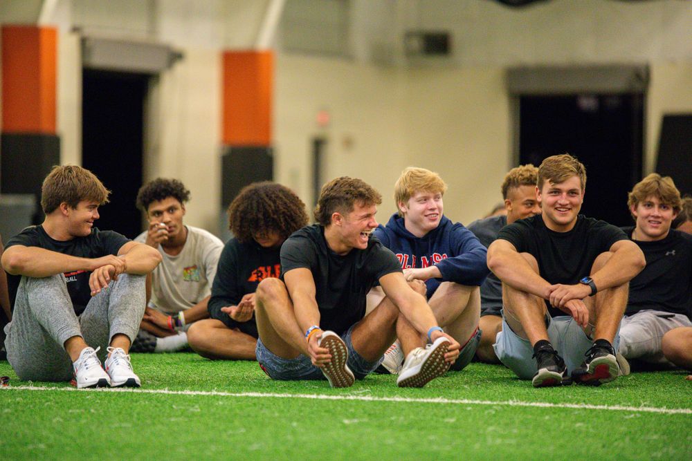 Row of people lying on green turf field during group activity indoors.