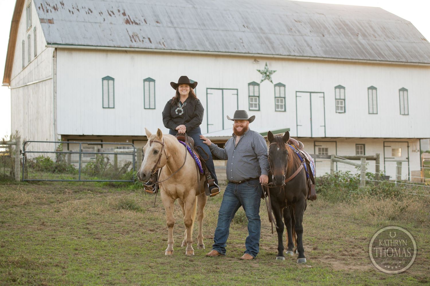Two horses and riders stand in front of a large white barn during golden hour.