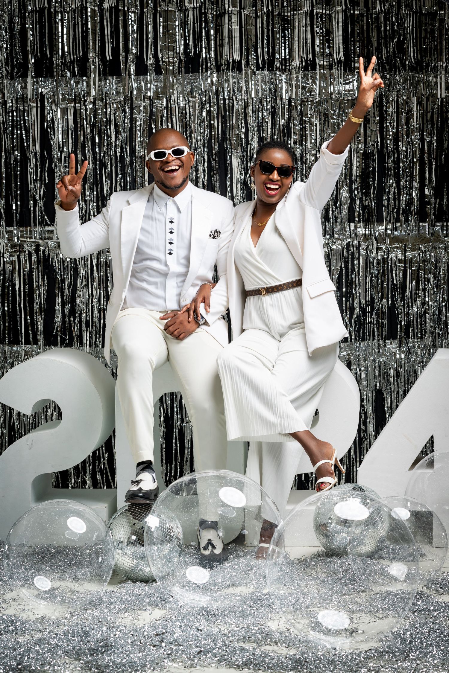 Stylish duo in matching all-white outfits pose with peace signs against a silver tinsel backdrop with 2024 numbers.