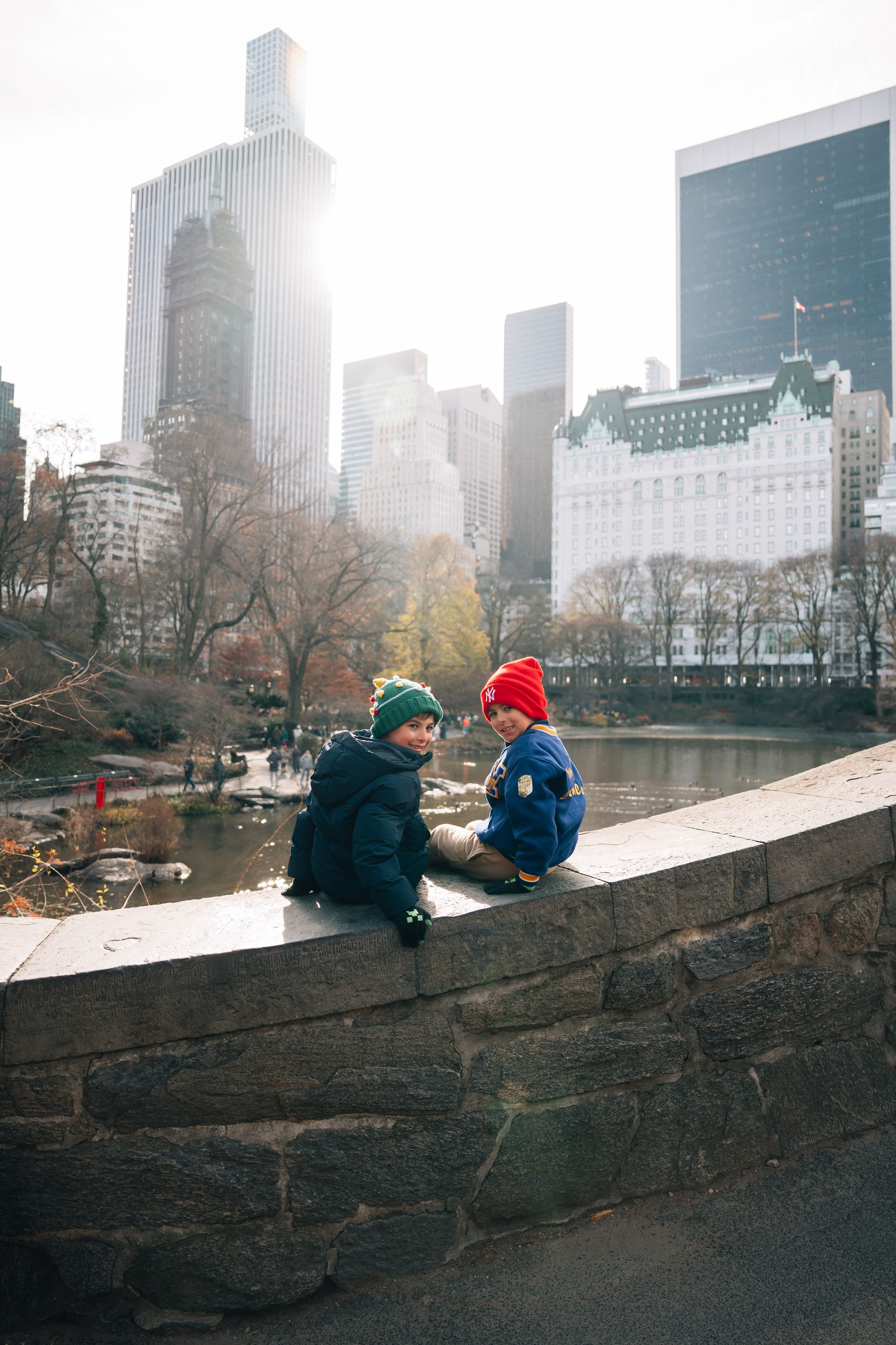 Two bundled up figures in winter hats sit together on a stone wall overlooking Central Park with NYC skyscrapers in background.