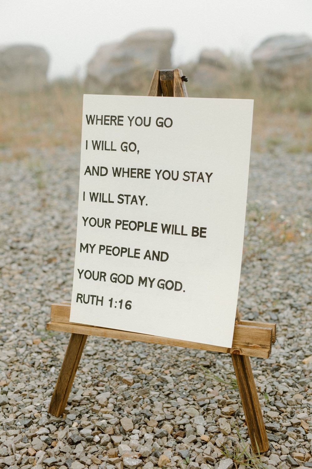 A wooden easel displays a sign with Ruth 1:16 Bible verse at an outdoor wedding ceremony.