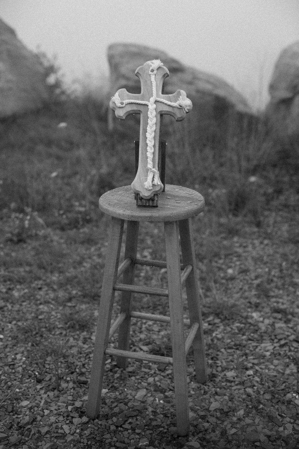 A decorative white cross rests on a wooden stool at an outdoor wedding ceremony in black and white.