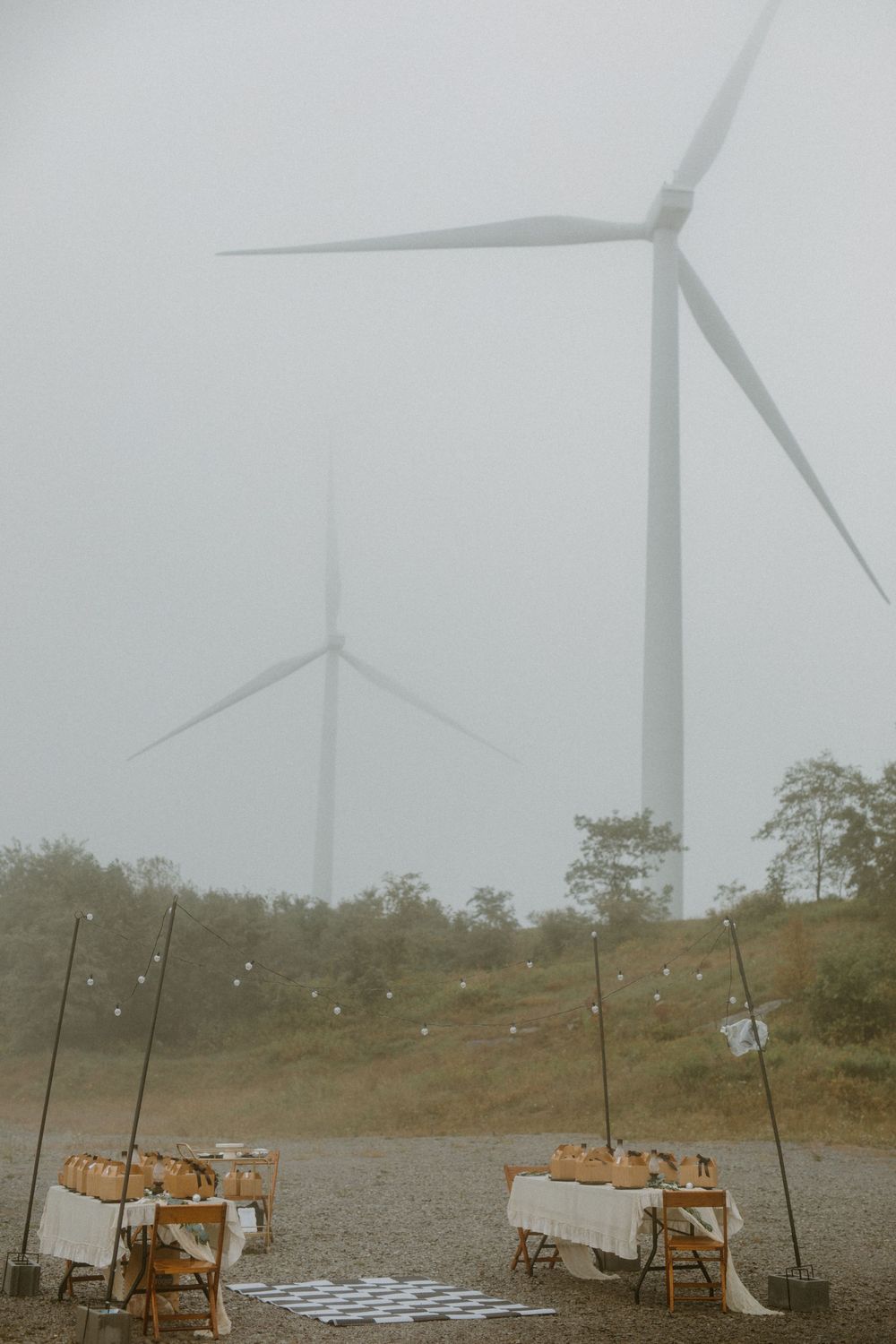 Two wind turbines loom in fog above rustic outdoor wedding reception tables on a misty day.