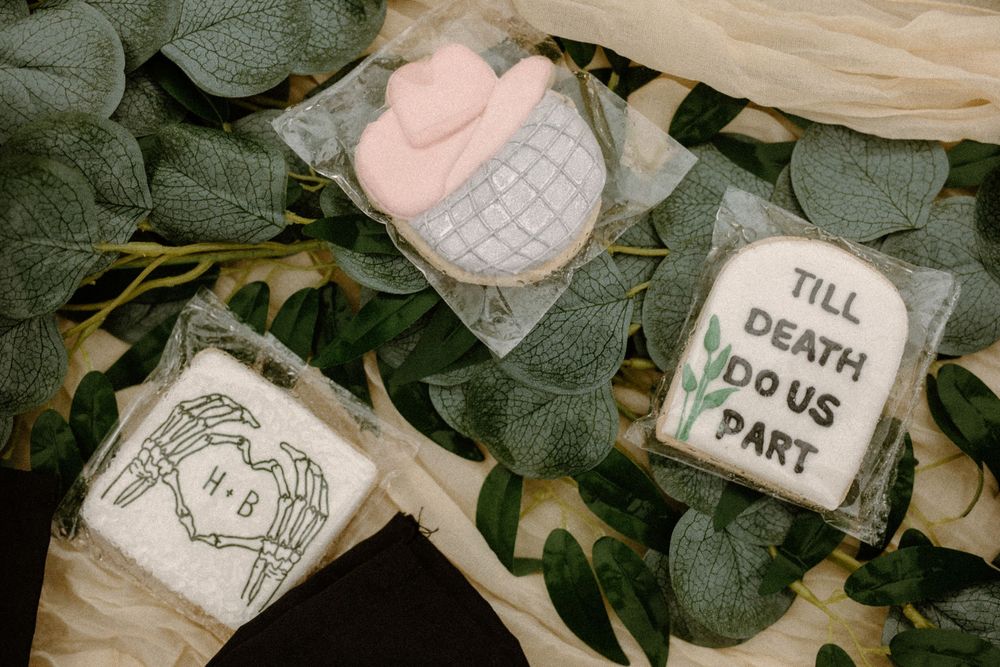 Wedding cookies decorated with a tombstone, skeleton hands, and pink hat design on green leaves.