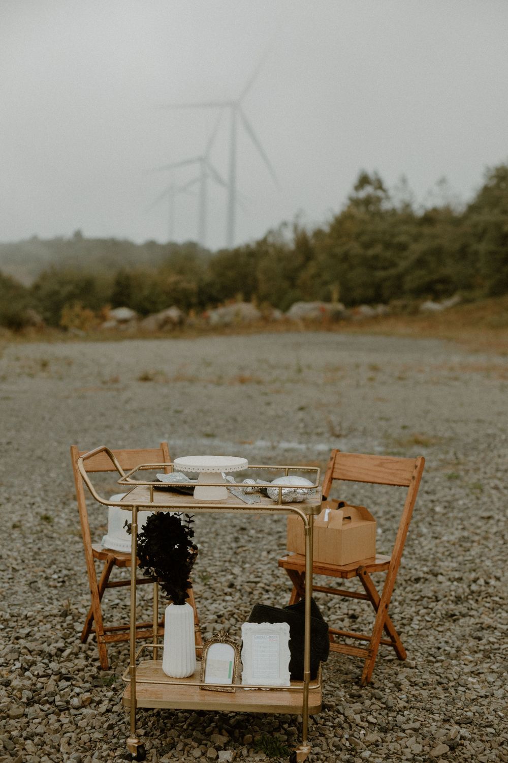 Rustic outdoor wedding table setup with wooden chairs and cart on gravel, wind turbines visible in misty background.