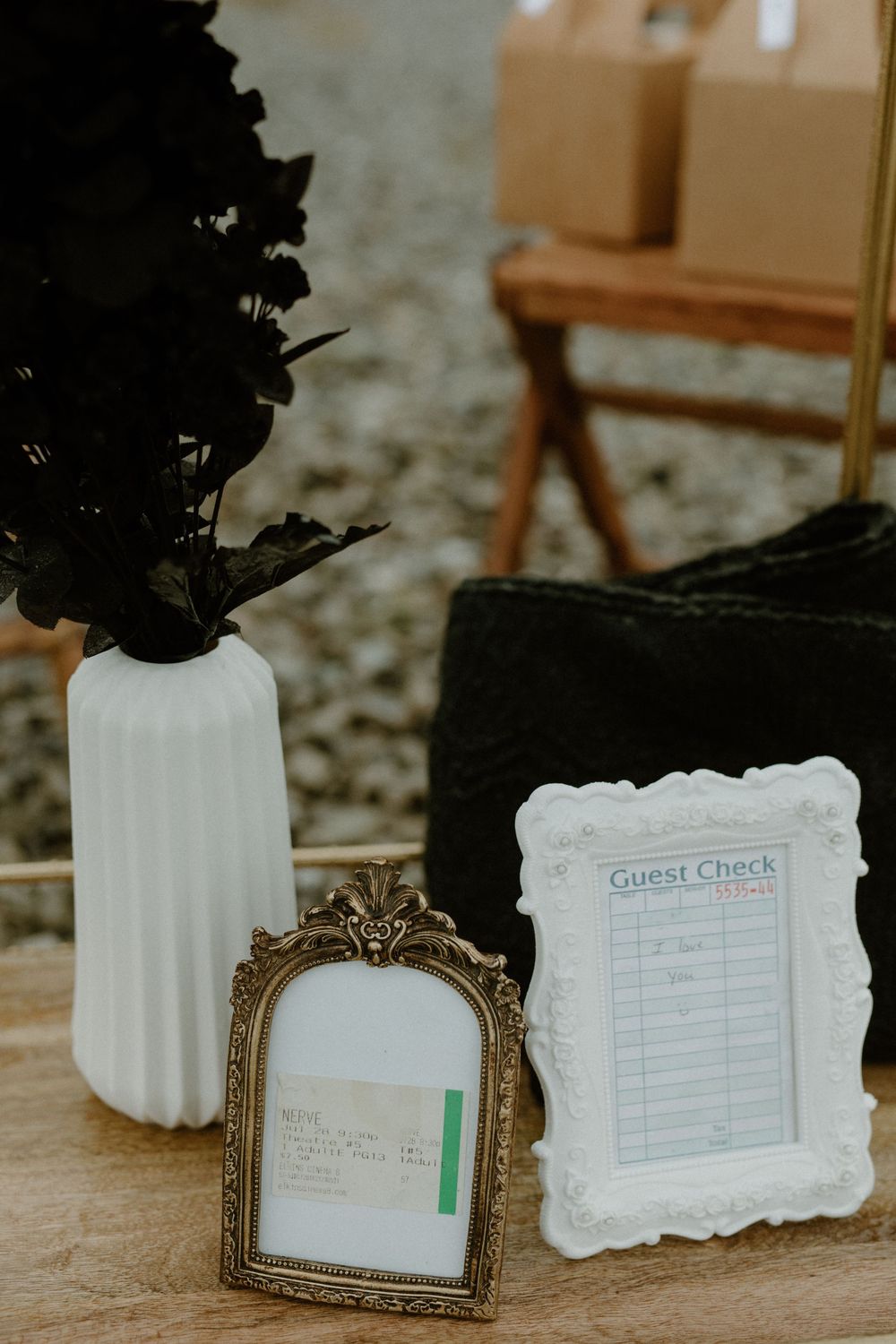 Elegant wedding guest check display with ornate gold frame, white ribbed vase holding dark florals on wooden surface.