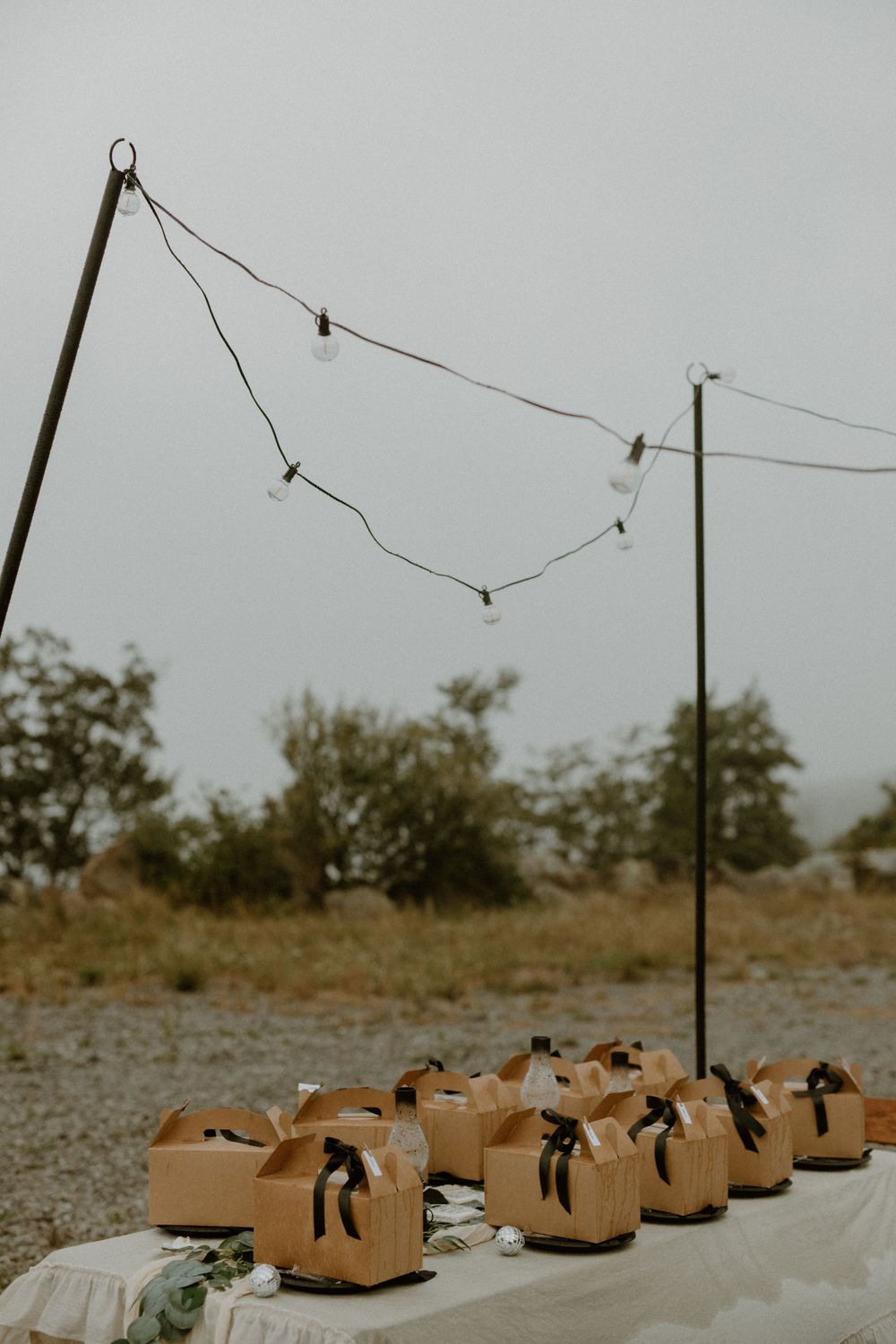 Kraft paper wedding favor boxes with black ribbon bows displayed on table under string lights on overcast day.