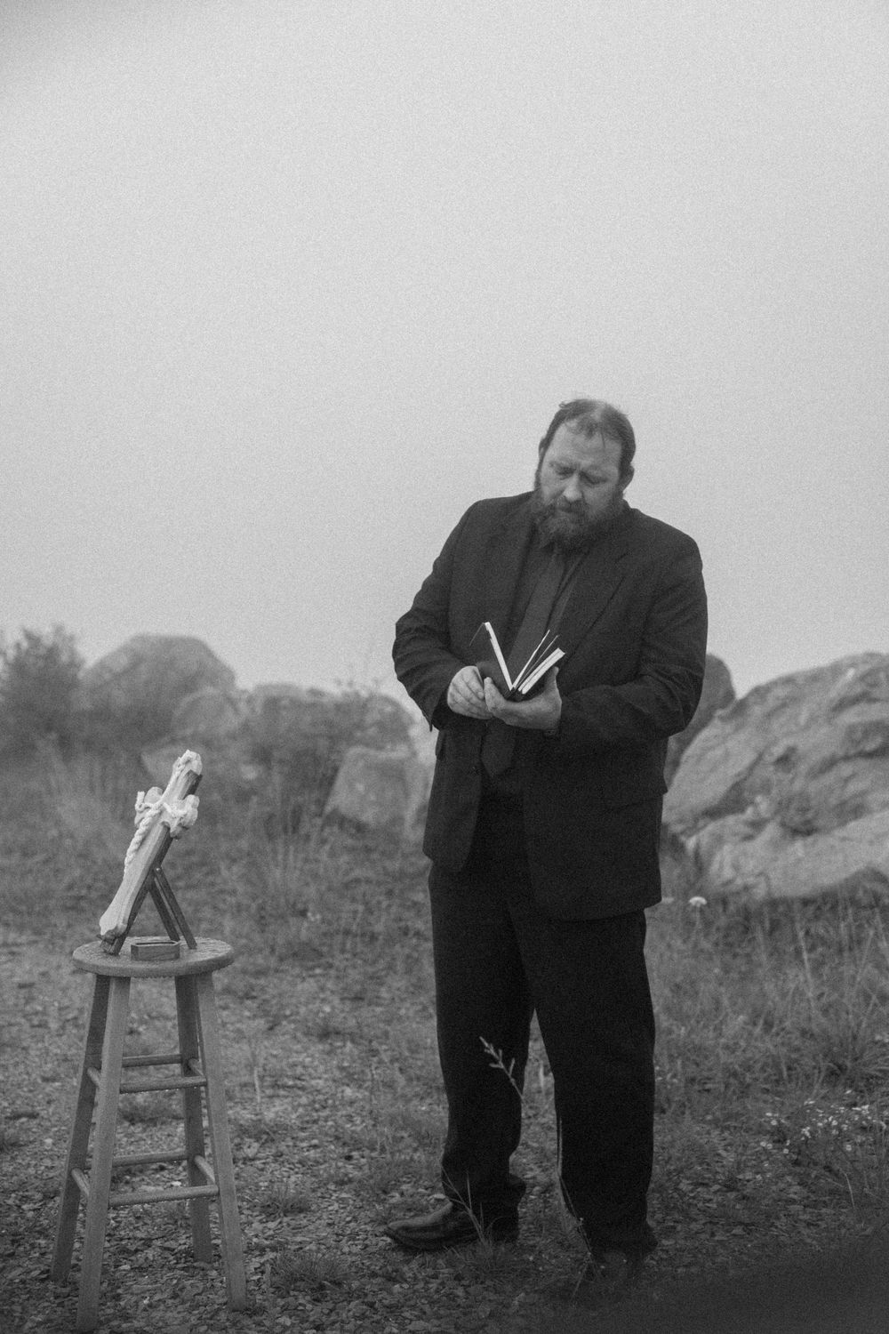 A bearded officiant in a black suit reads from a book at an outdoor wedding ceremony in foggy weather.