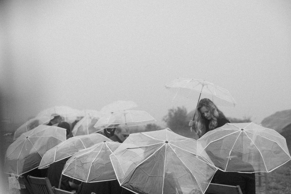 Wedding guests holding clear umbrellas in the rain at an outdoor ceremony captured in black and white.