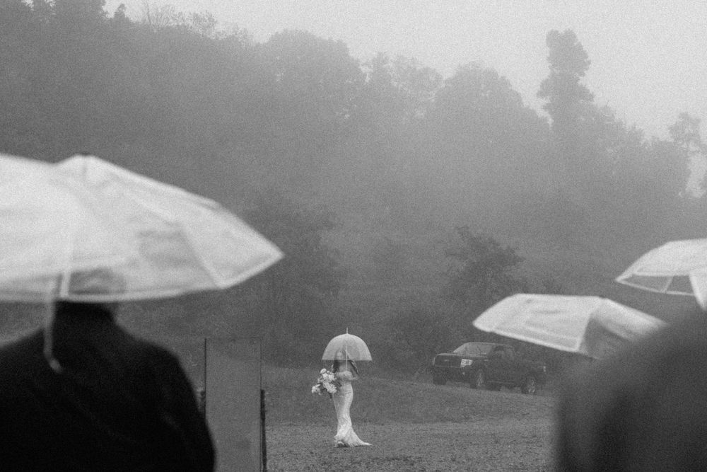 A bride carrying an umbrella and bouquet walks through a foggy rainy outdoor wedding venue.