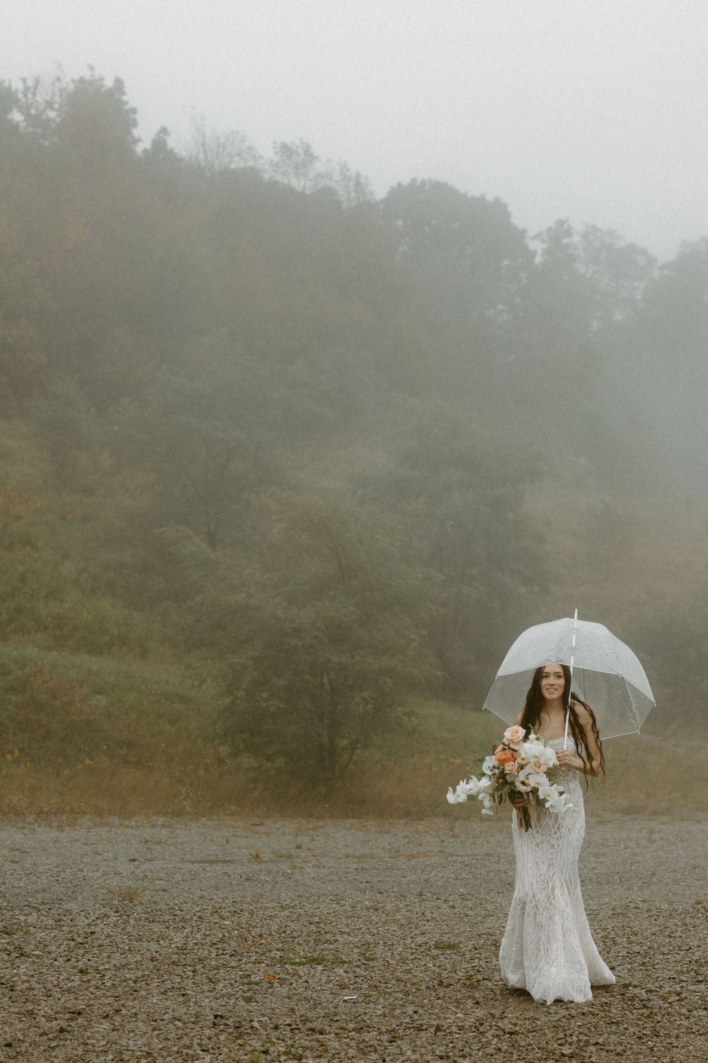 A bride in a lace gown holds a clear umbrella and bouquet in a misty foggy outdoor setting.