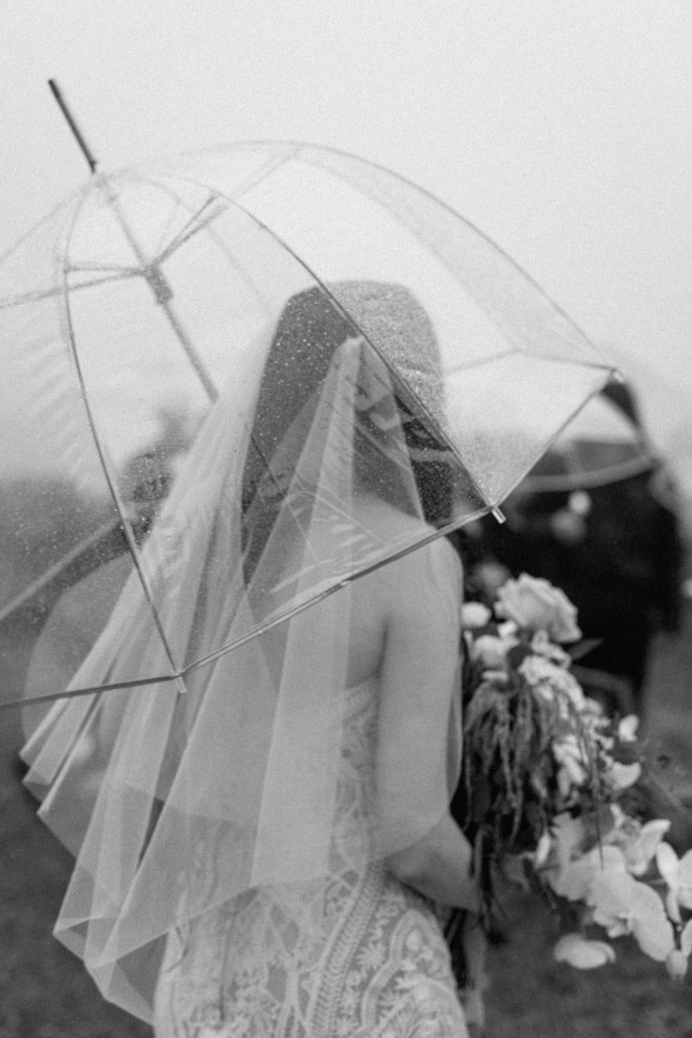 Black and white photo of bride in lace gown and veil holding clear bubble umbrella on rainy wedding day.