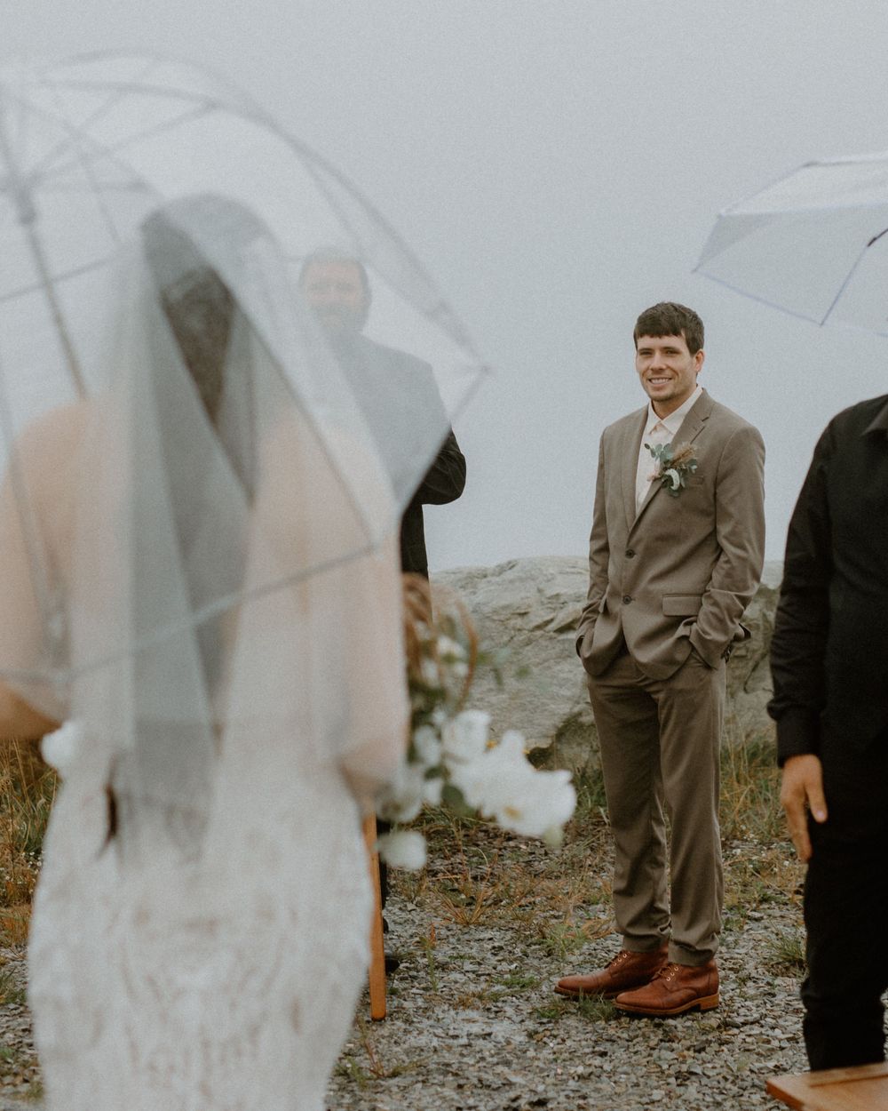 Groom in grey suit smiling at veiled bride approaching during outdoor rainy wedding ceremony with umbrellas.