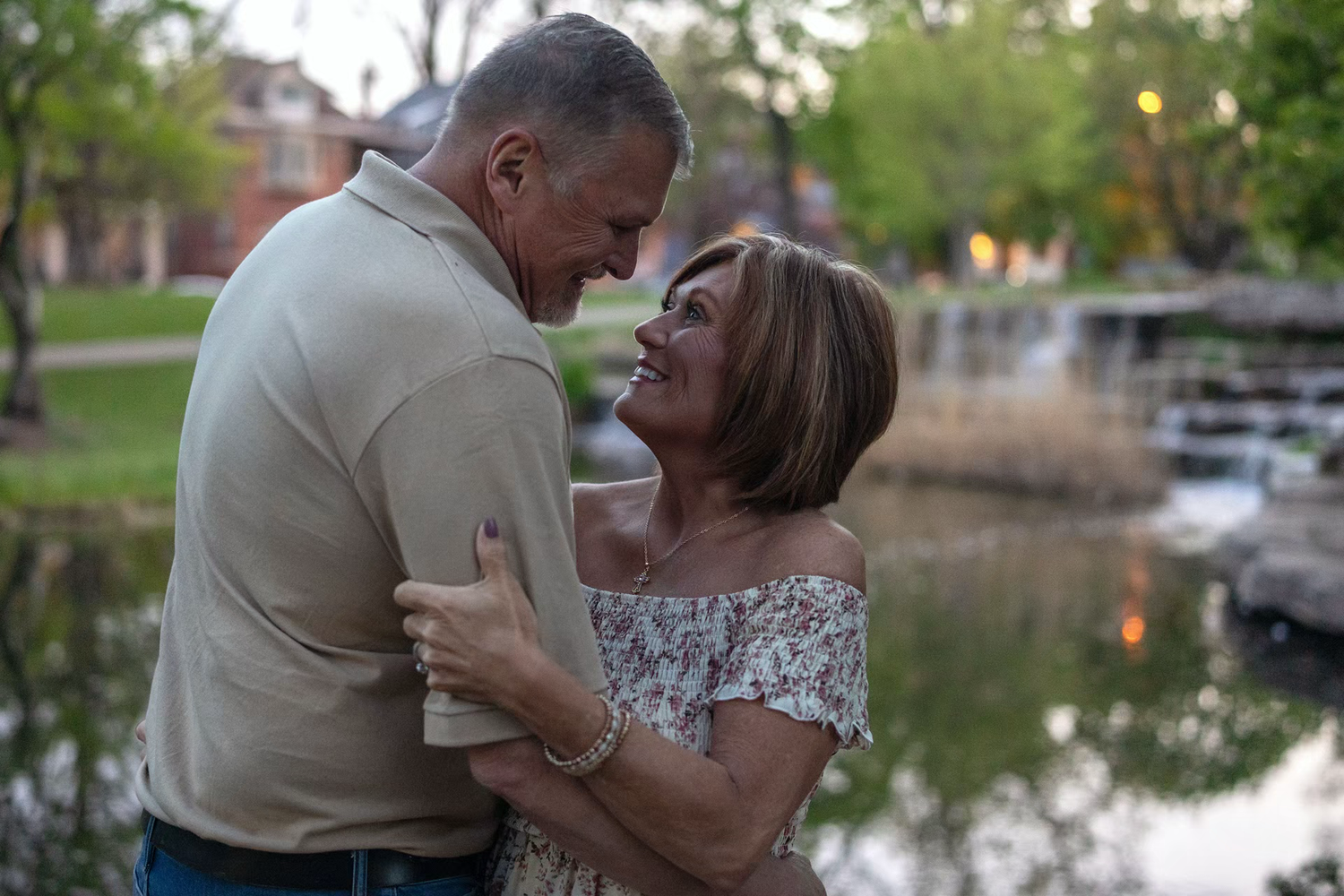 Older couple embracing and smiling at each other near a pond in a lush green park at dusk.