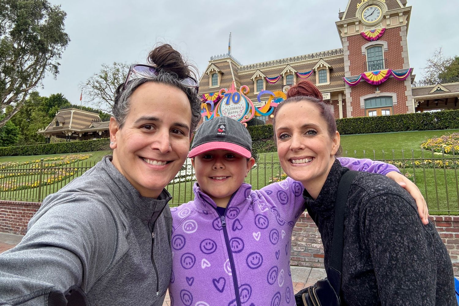 The Mamones smiling for a selfie at Disneyland entrance on a cloudy day, wearing casual athletic wear because they're not working