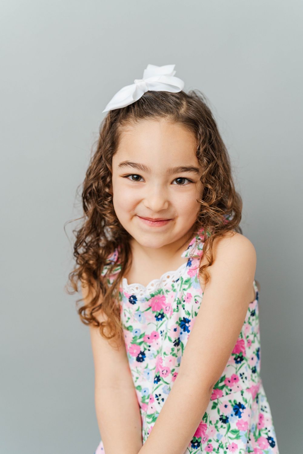 Young girl with curly hair and white bow wearing a floral dress, smiling against a gray background.