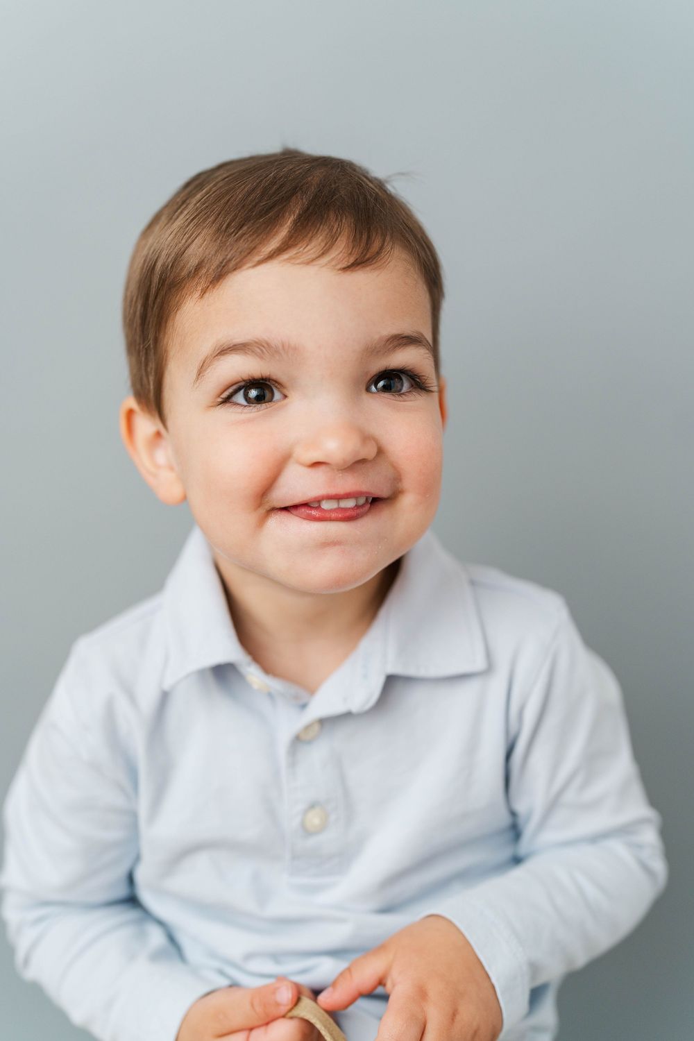 Adorable toddler boy in a light blue polo shirt smiling cheerfully against a gray background.
